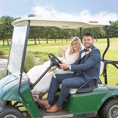 A couple sat on a golf buggy with lush green trees behind them.