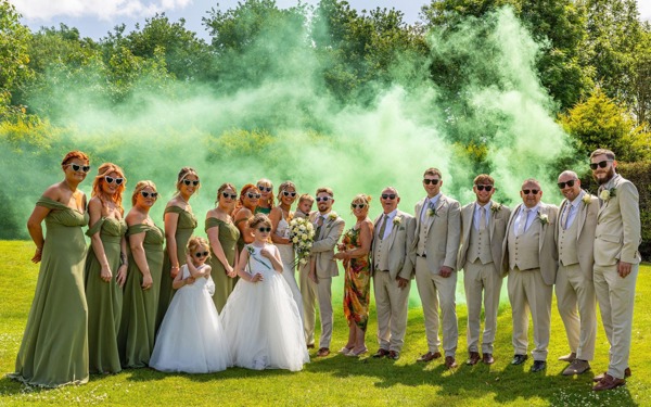 A bride and groom holding their young son are flanked by their bridesmaids and groomsmen on The Lawn of  The Manor; green smoke billows behind them.