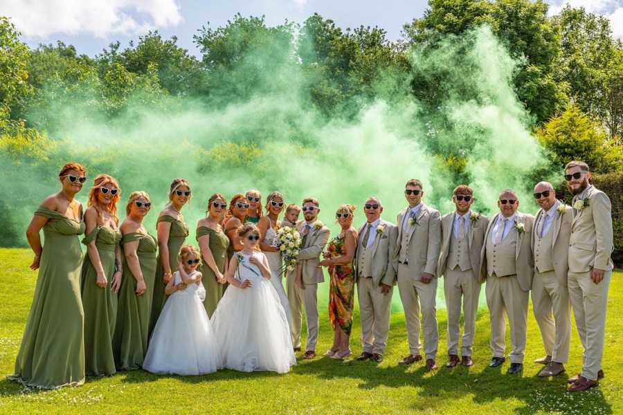 A bride and groom holding their young son are flanked by their bridesmaids and groomsmen on The Lawn of  The Manor; green smoke billows behind them.
