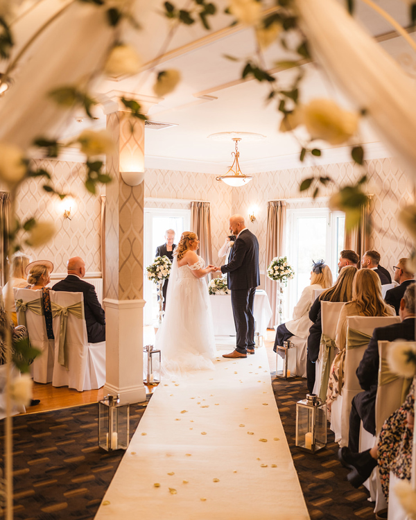 A wedding ceremony taking place in High Garden at The Manor. The view is through a floral archway, the bride and groom face each other at the end of the aisle.