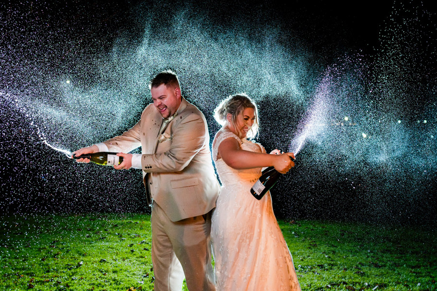 A night shot of a bride and groom spraying prosecco on The Lawn of The Manor.