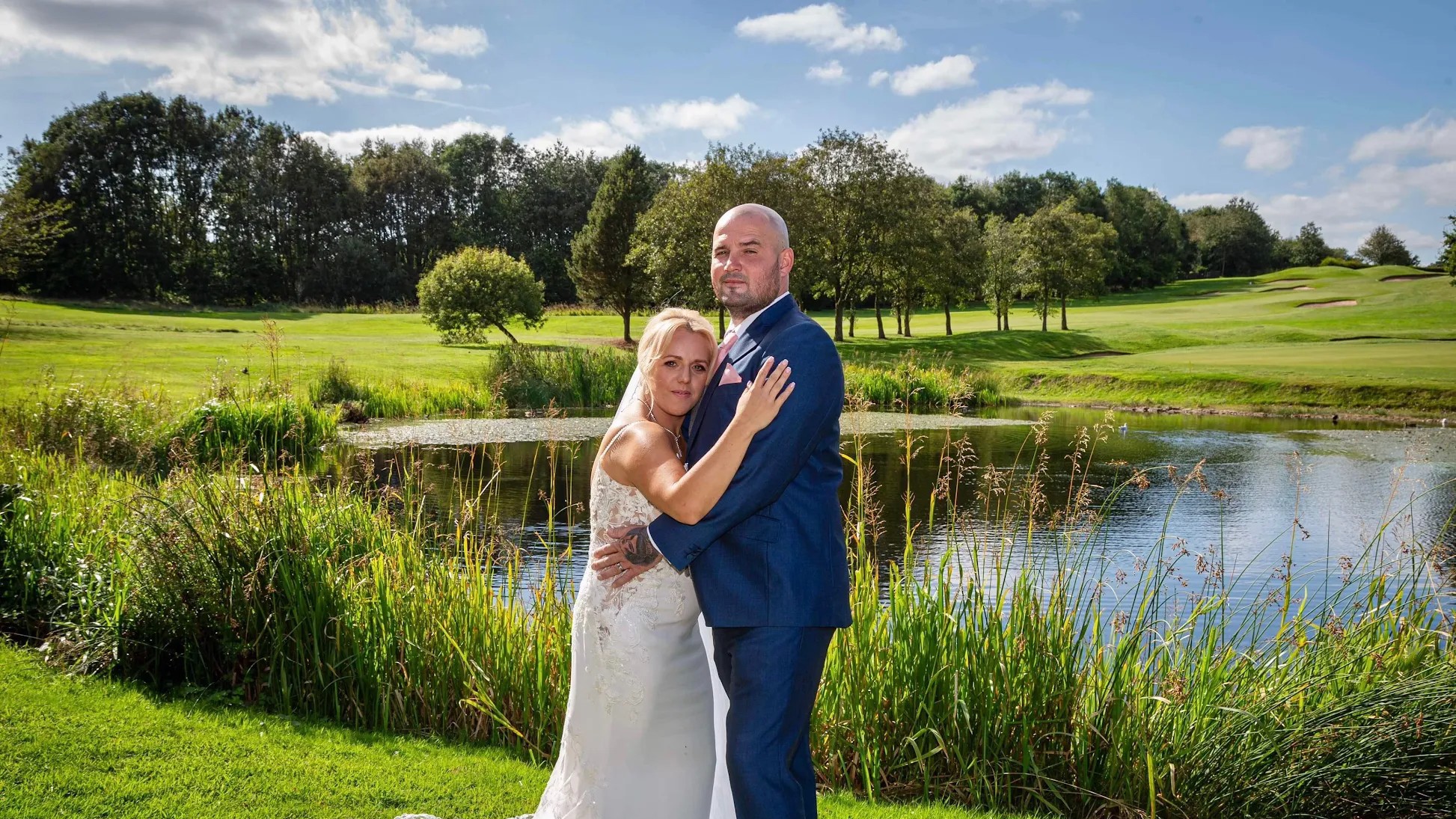A bride and groom embrace in front of The Lake at The Manor Leeds on a sunny day.