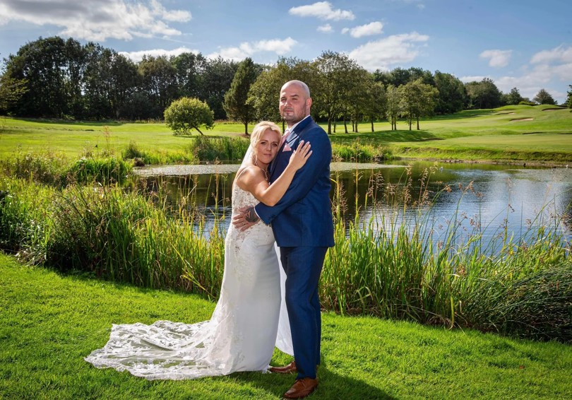 A bride and groom embrace in front of The Lake at The Manor Leeds on a sunny day.