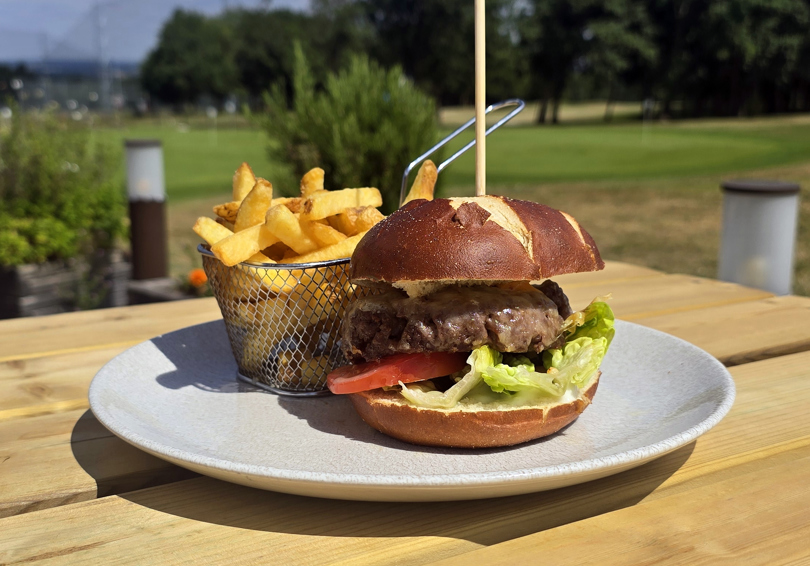 A beef burger in a pretzel bun with lettuce and tomato and a wire basket of fries sitting on a plate placed on an outside wooden table. There are trees in the background and the netting of The Manor Leeds's driving range can be seen.