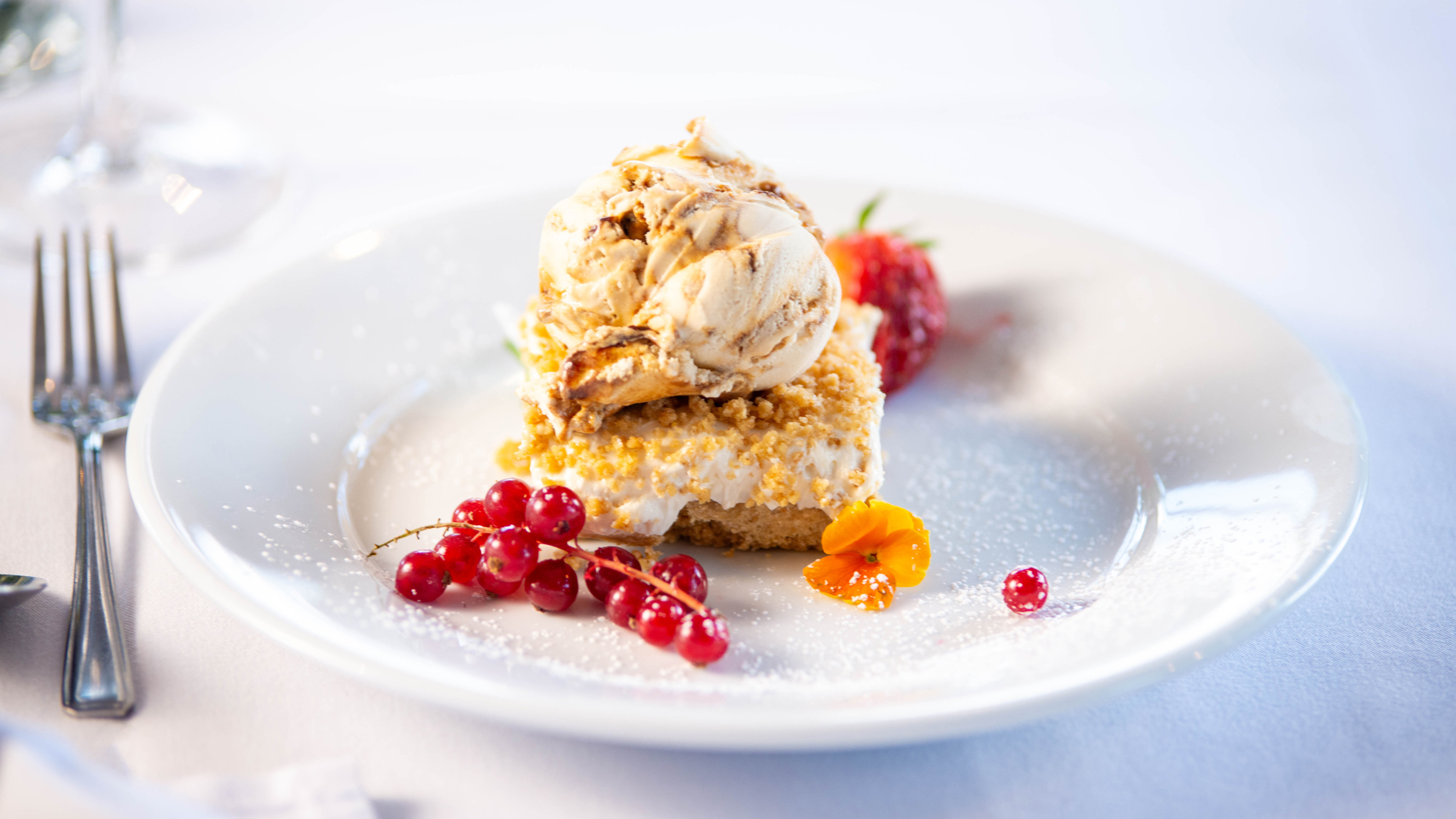Cheesecake on a plate, topped with ice cream, red currents and an edible flower sit beside.