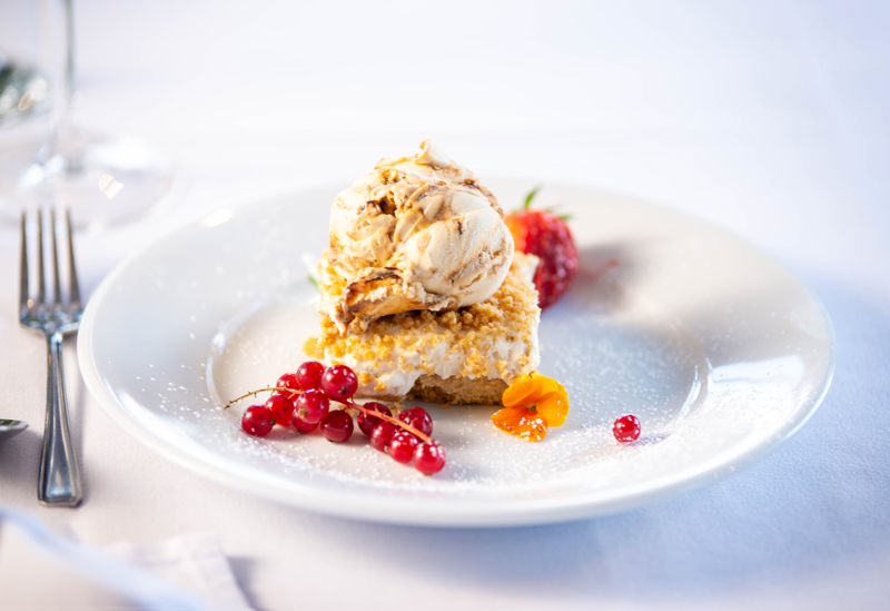 Cheesecake on a plate, topped with ice cream, red currents and an edible flower sit beside.