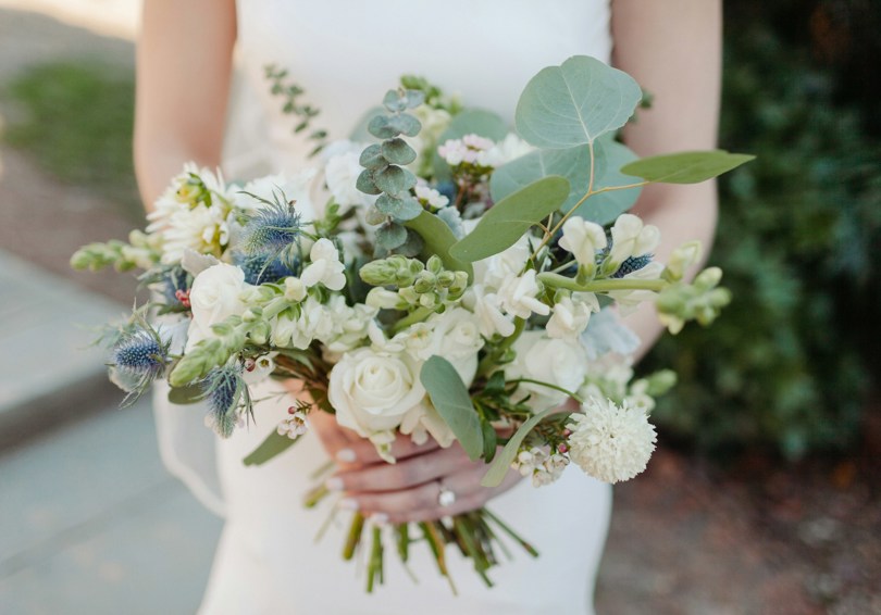 A bouquet of flowers held by a bride, featuring eucalyptus, thistles, stocks and white roses.