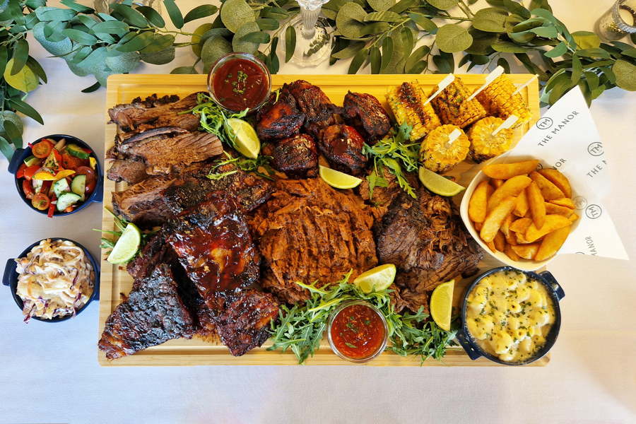 The Grill sharing board at The Manor Leeds; Slow-cooked BBQ Beef Brisket, Sticky BBQ Chicken Thighs, Pulled Pork with Smoky Glaze, Pork Ribs, Corn on the Cob, Mac & Cheese, Skin-on Wedges, Classic Crunch Slaw and Tomato & Cucumber Salad.