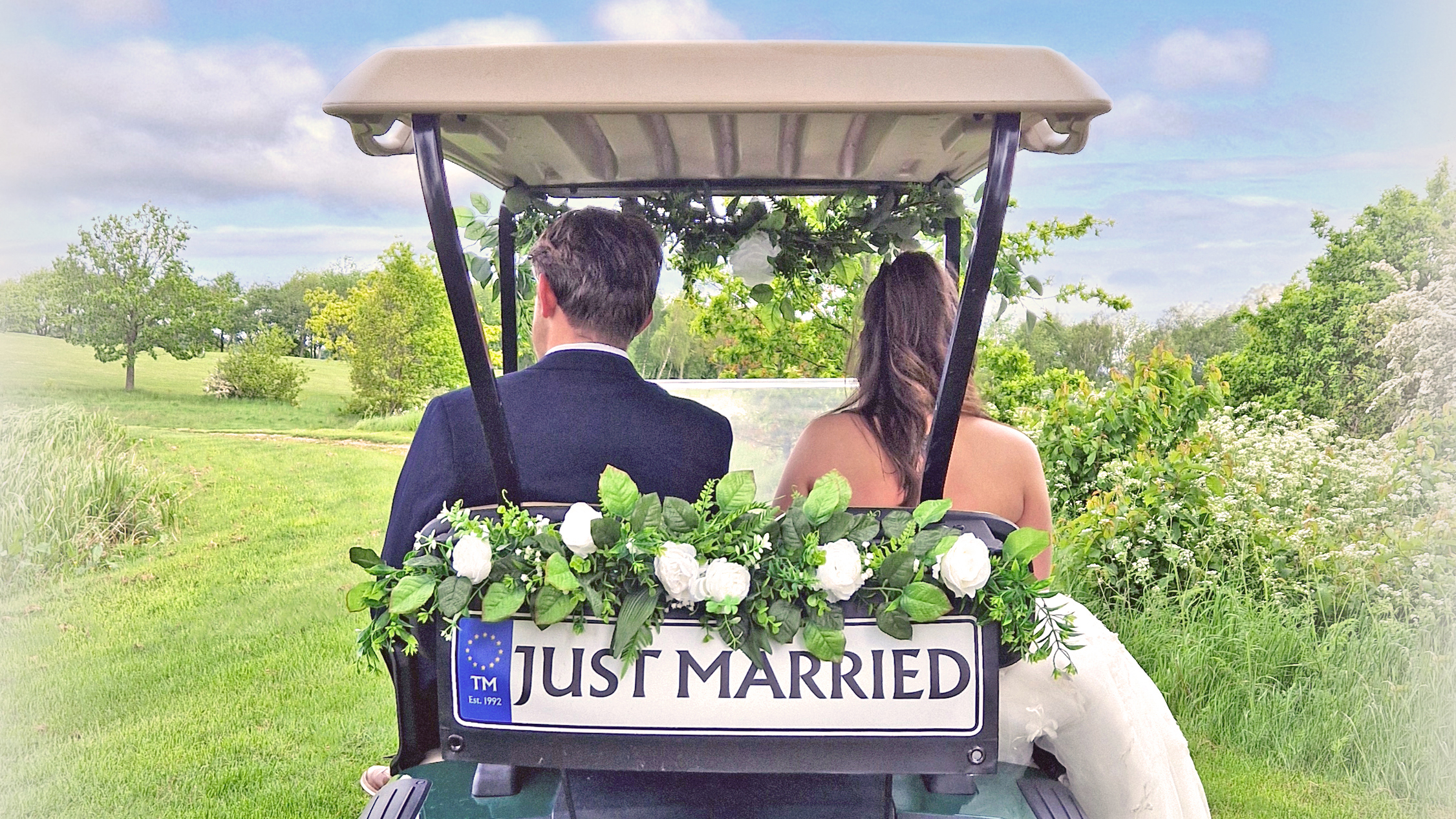 A rear view of a bride and groom sitting in a golf buggy decorated with a "Just Married" sign and white flowers.