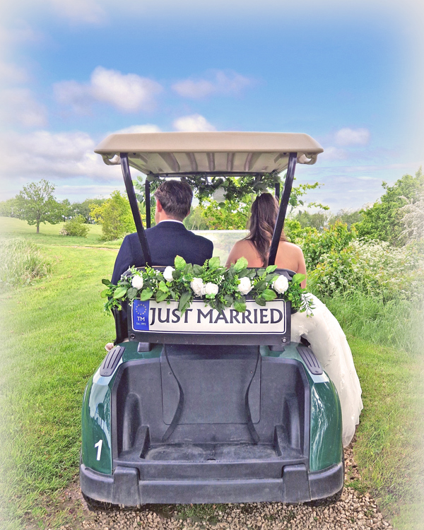 A rear view of a bride and groom sitting in a golf buggy decorated with a "Just Married" sign and white flowers.