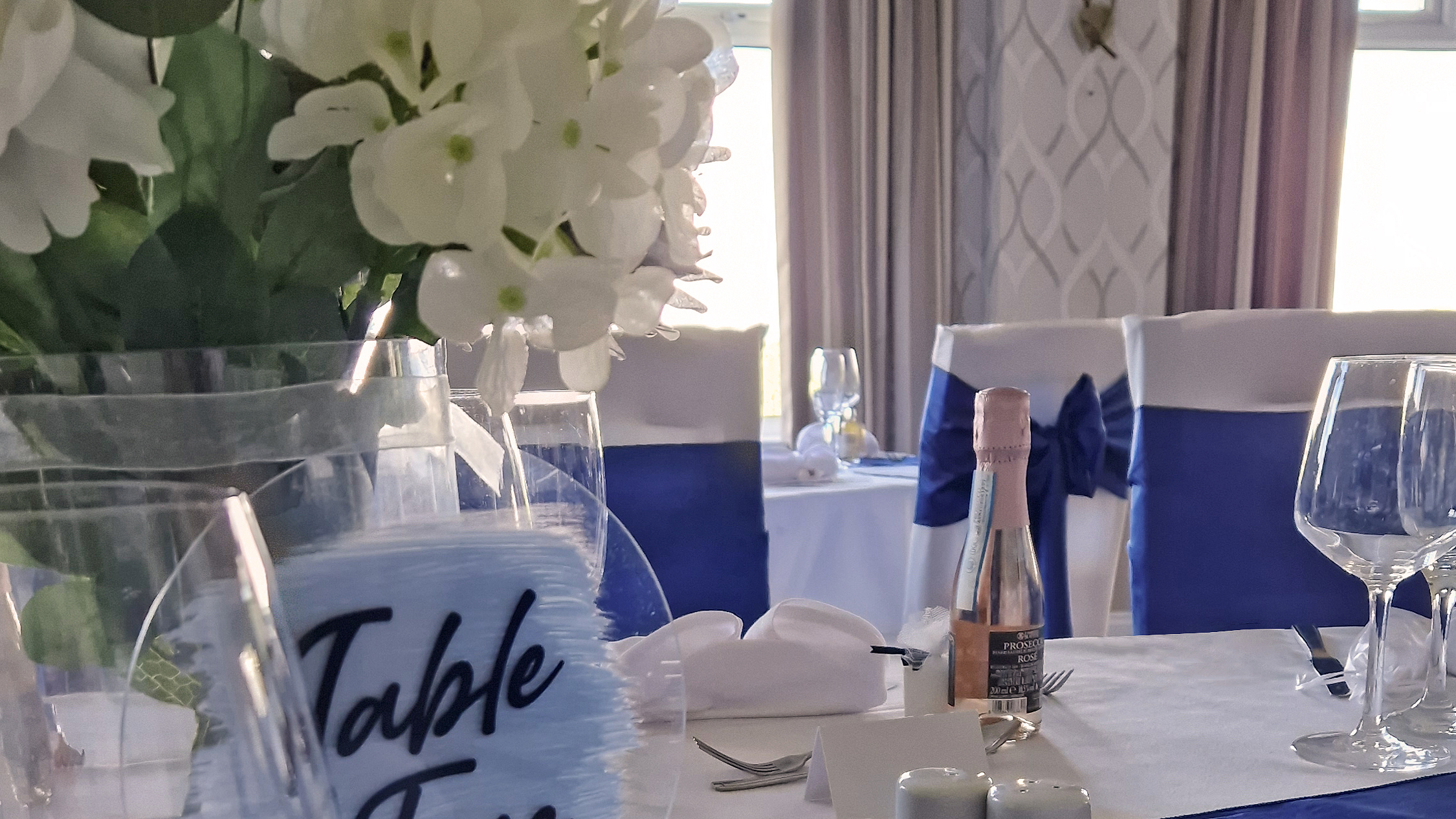 A dining table at The Manor set for a wedding breakfast, the table runner and chair sashes are royal blue, white hydrangeas sit in a vase on a wood slice in front of a sign saying "Table Five".