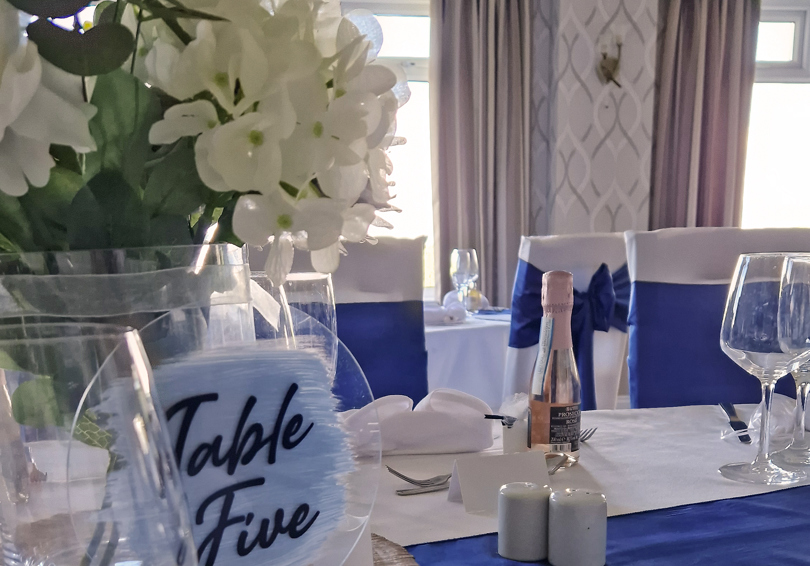 A dining table at The Manor set for a wedding breakfast, the table runner and chair sashes are royal blue, white hydrangeas sit in a vase on a wood slice in front of a sign saying "Table Five".