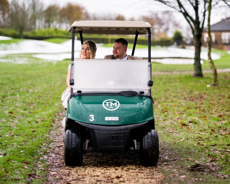 A bride and groom in a green golf buggy featuring The Manor's logo.