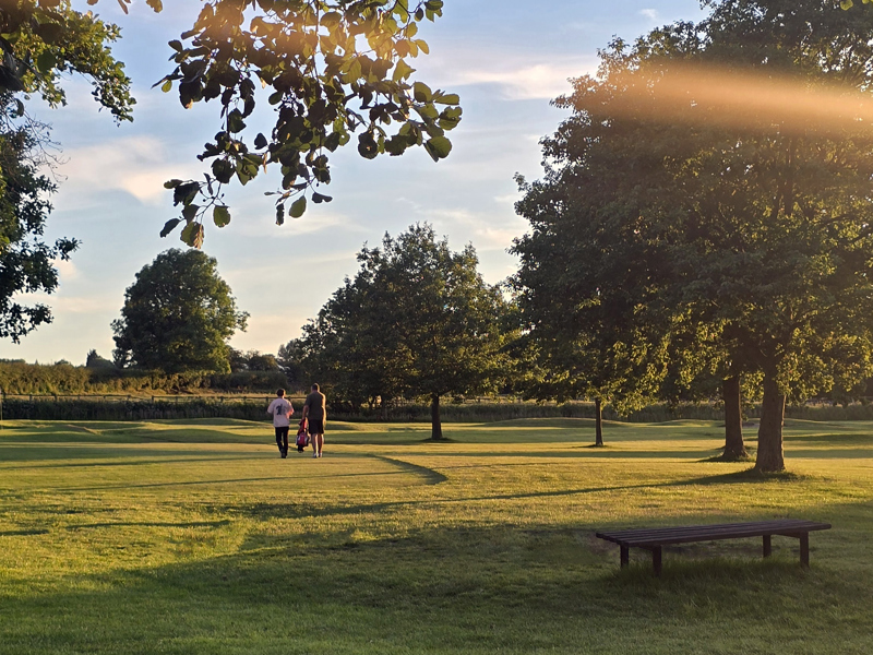 View of the Par 3 Academy Course at The Manor Leeds taken on a summer evening, a man and adolescent boy are walking away with a golf trolley between them.