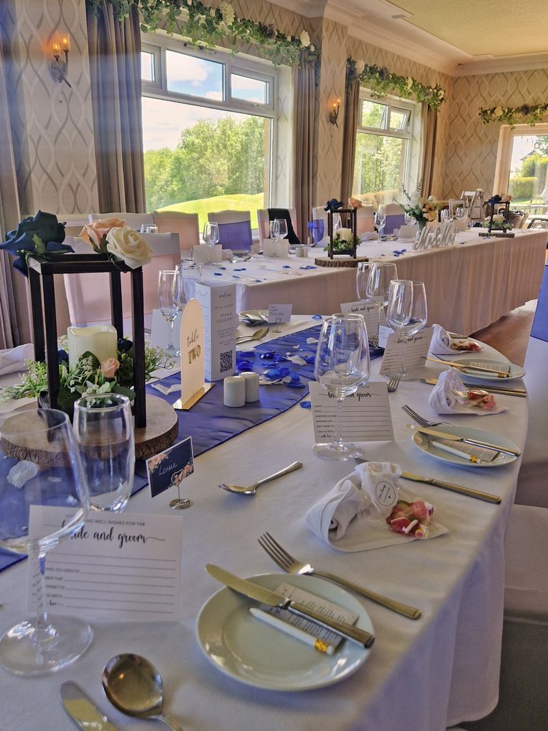 A dining table at The Manor set for a wedding breakfast, the white linen napkins are folded into hearts and the table runner is dark blue. The long top table can be seen in the background and grass and trees can be seen through the windows.