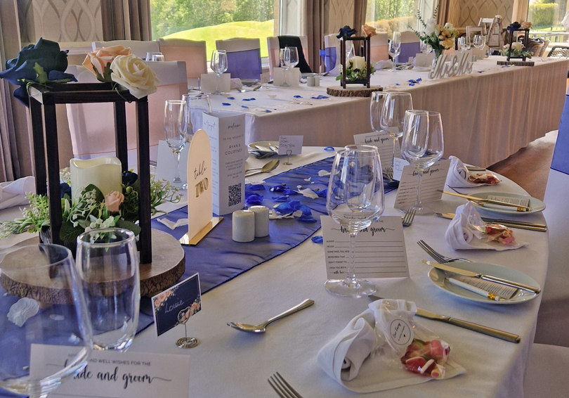 A dining table at The Manor set for a wedding breakfast, the white linen napkins are folded into hearts and the table runner is dark blue. The long top table can be seen in the background and grass and trees can be seen through the windows.