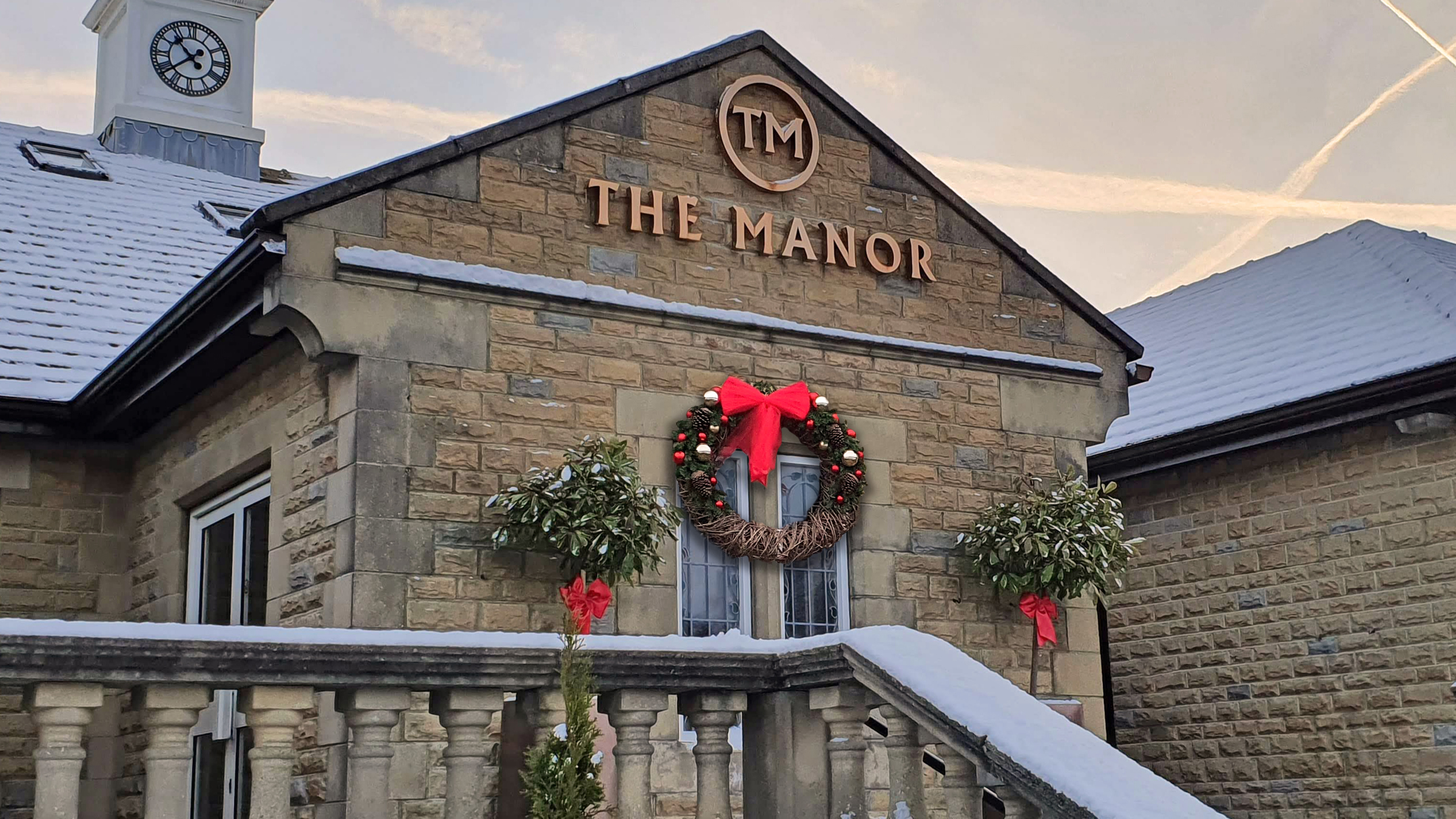 The main entrance of The Manor, showing the stone balustrade, the clocktower and the gold logo, a large Christmas wreath with a red bow hangs beneath the sign.