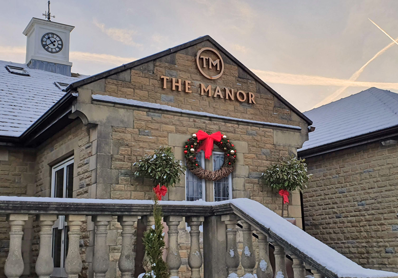 The main entrance of The Manor, showing the stone balustrade, the clocktower and the gold logo, a large Christmas wreath with a red bow hangs beneath the sign.
