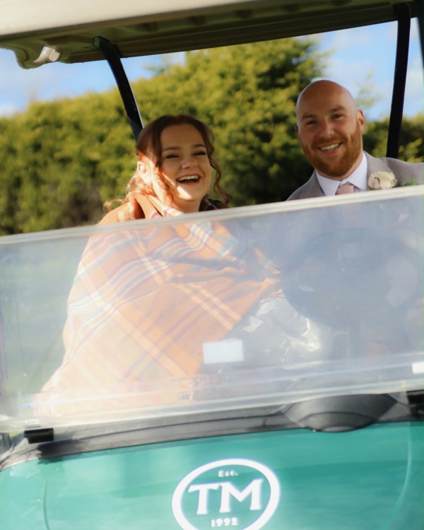 A bride and groom smiling from a green golf buggy featuring The Manor's logo.