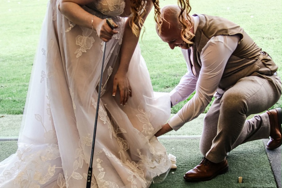 A bride and groom in The Manor's driving range, the bride is holding a golf club, the groom is on one knee adjusting her dress.