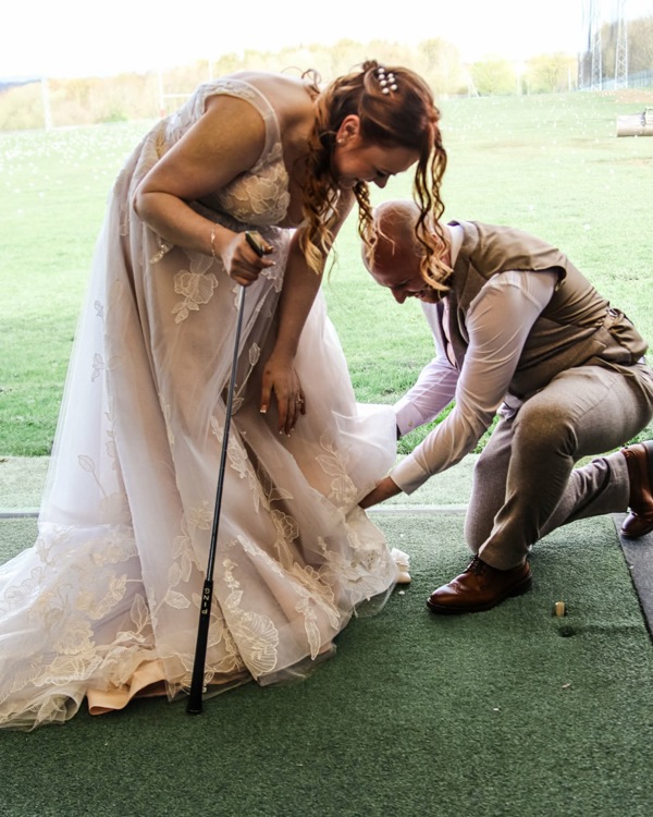 A bride and groom in The Manor's driving range, the bride is holding a golf club, the groom is on one knee adjusting her dress.