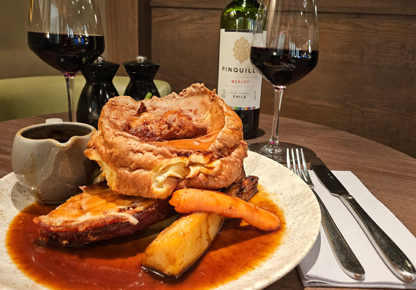 A photo of a roast diner on a plate, including a large Yorkshire pudding, a roasted carrot and parsnip, and a small rug of gravy. A knife and fork lay on a folded napkin to the right hand side and two glasses of red wine can be seen in the background along with the bottle and salt and pepper mills.