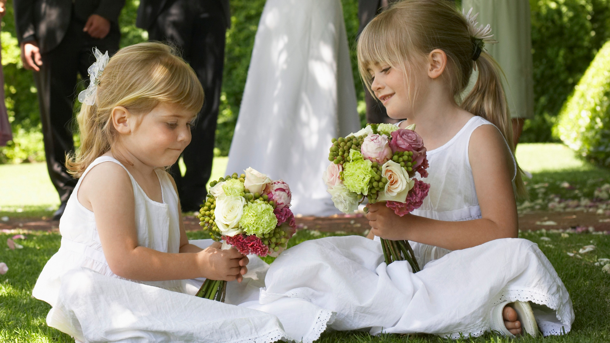 Two young bridesmaids in white dresses sitting on grass down admiring their flower posies.