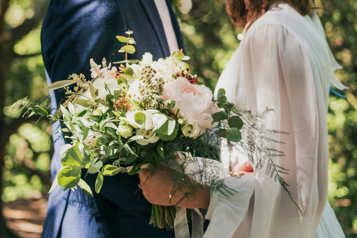 A headless shot of a bride and groom, facing each other. The bride is holding a large, loose bouquet that features pale pink peonies, white roses and eucalyptus.