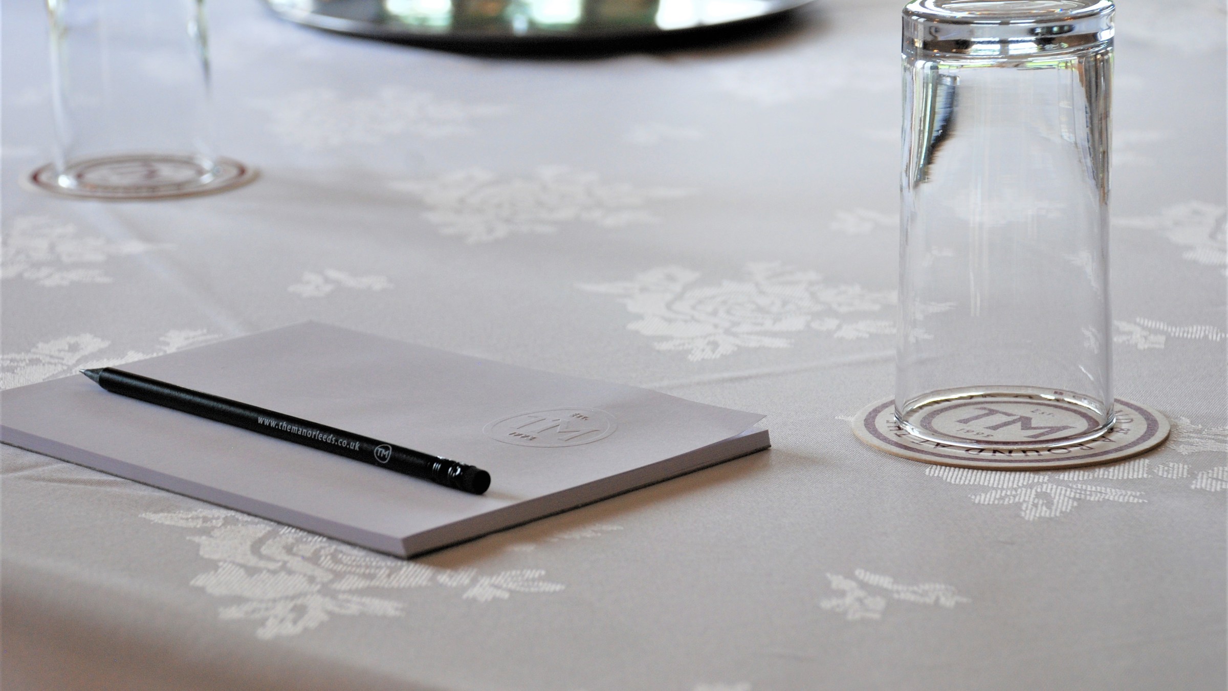 A notebook, pencil and upturned water glass placed on a table covered with a white cloth.