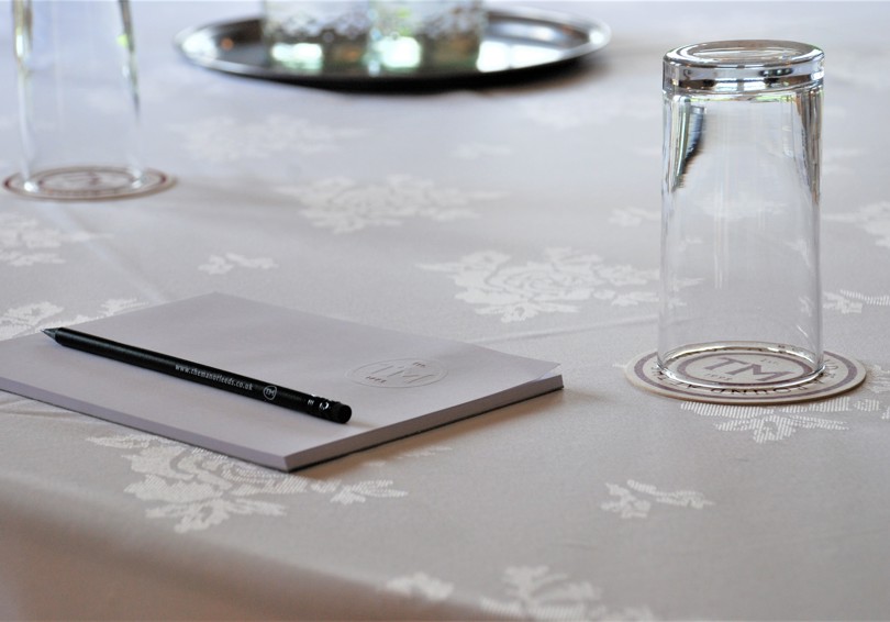 A notebook, pencil and upturned water glass placed on a table covered with a white cloth.