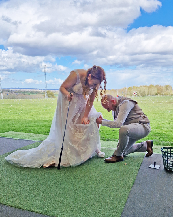 A bride and groom in The Manor's driving range, the bride is holding a golf club, the groom is on one knee retrieving a golf ball from beneath her dress.