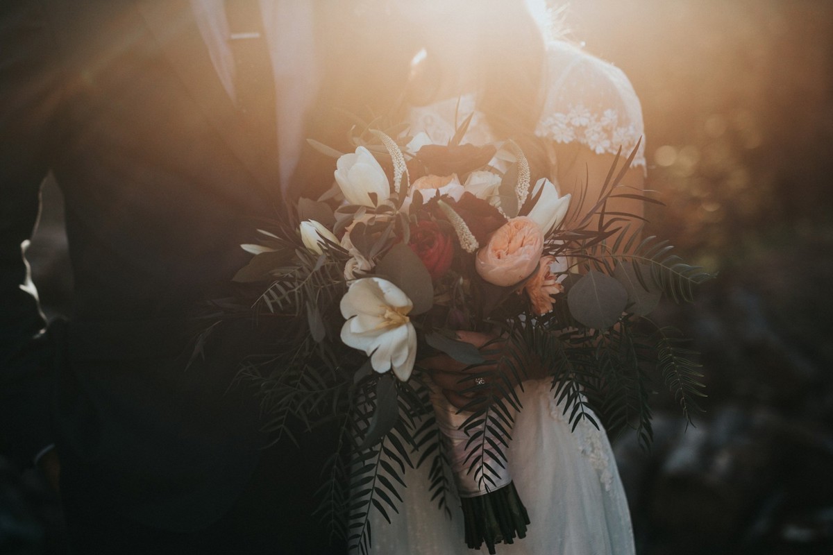 A headless shot of a bride and groom, taken at sunset. The bride is holding a bouquet that features peach-coloured peonies and palm fronds.